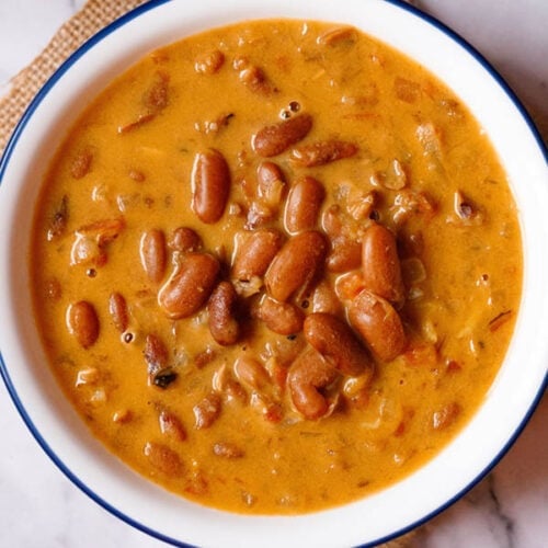 overhead shot of rajma recipe in a blue rimmed white bowl on a jute cloth with a brass spoon and bowl with onion rounds and lemon wedges