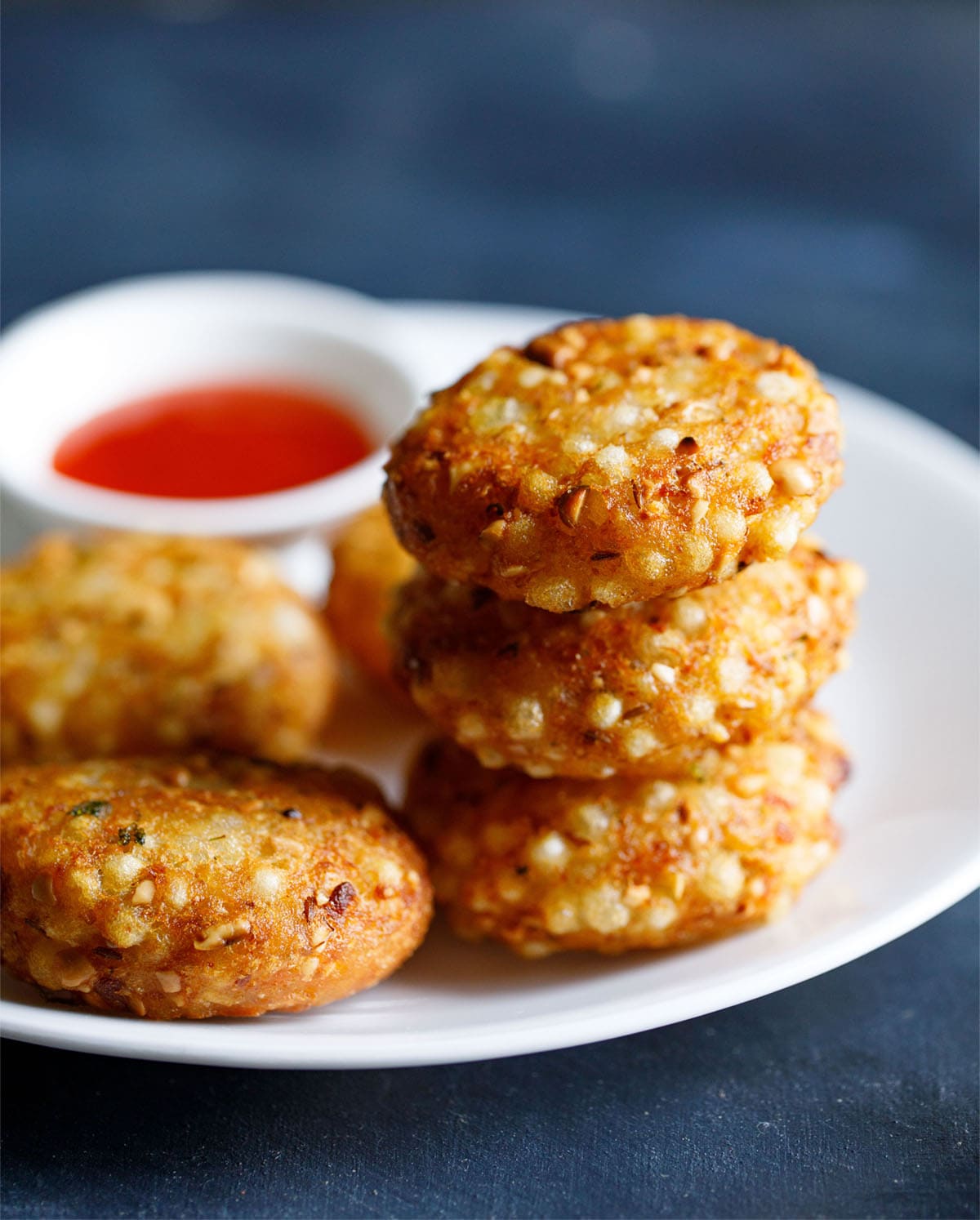 closeup shot of a stack of sabudana vada on top of each other in a white plate with red chilli sauce in a white bowl