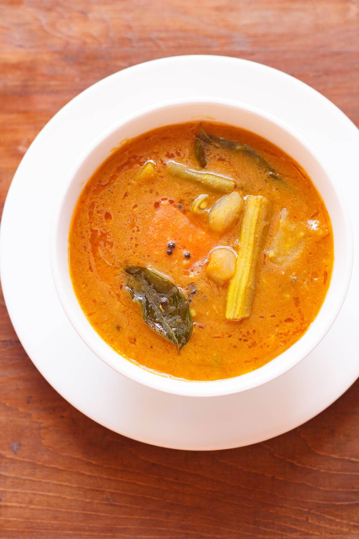 overhead shot of sambar filled in a white bowl placed on white plate on a brown wooden board