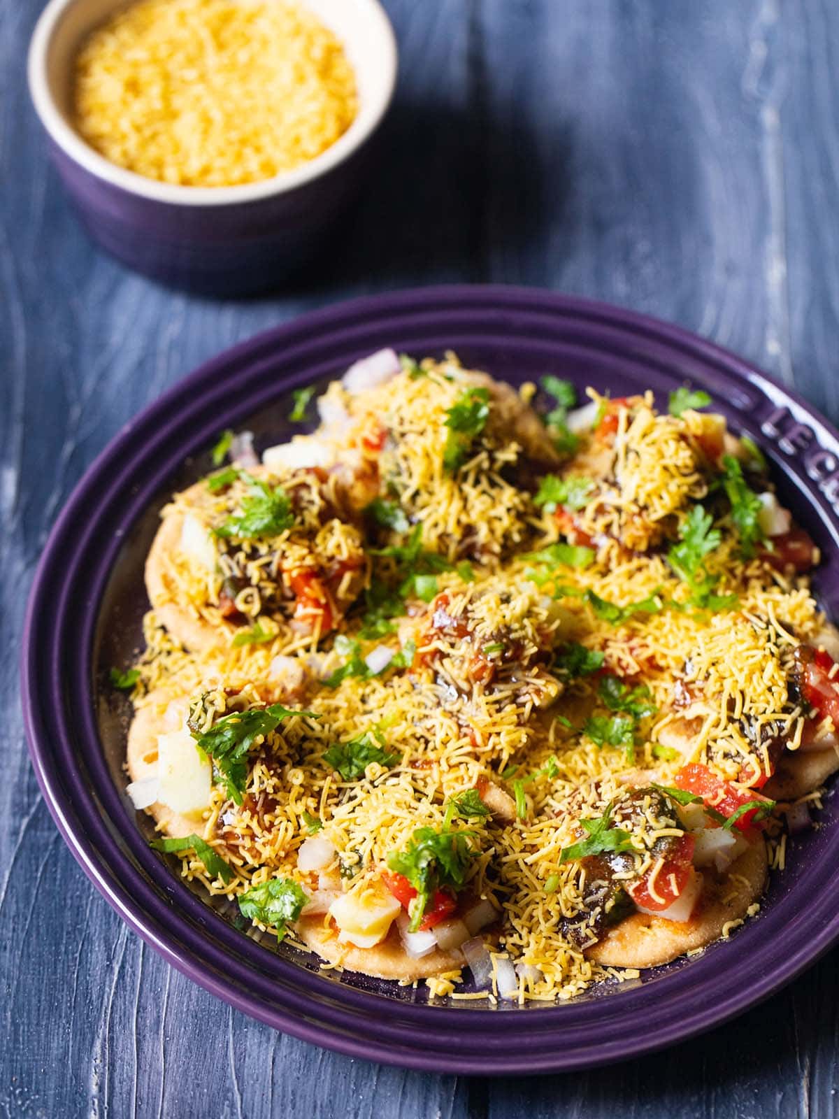 closeup shot of sev puri on a dark purple plate on a a dark blue table