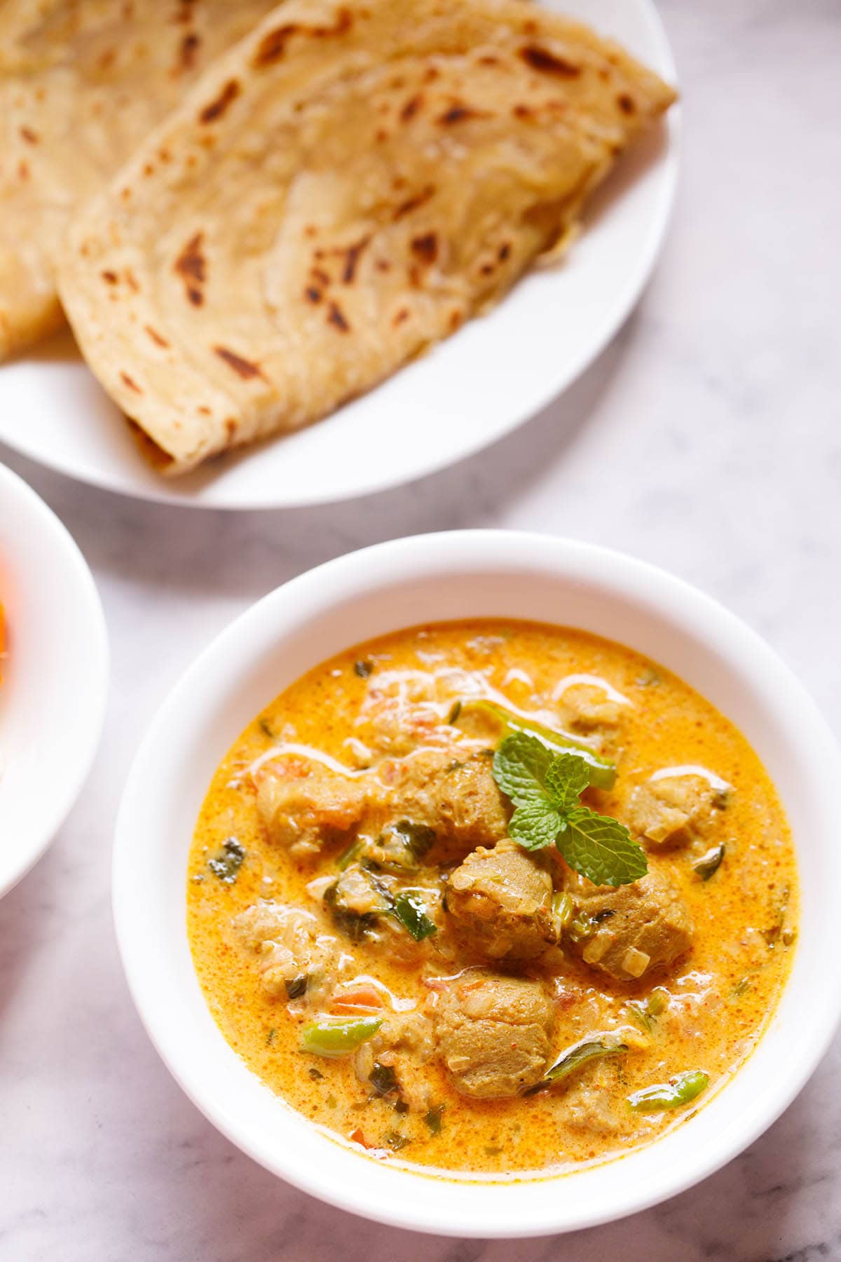 overhead shot of a white bowl of soya chunks curry on a white table with parathas in the background