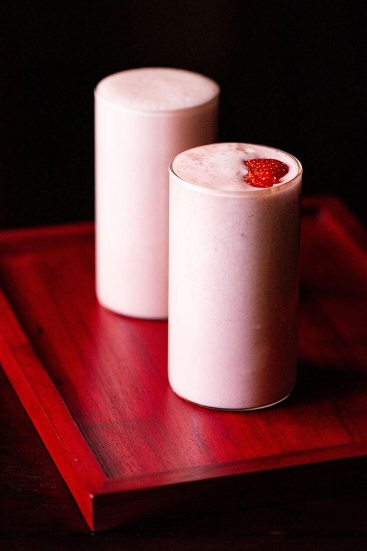 strawberry milkshake topped with strawberry slices in two glasses on a reddish pink tray