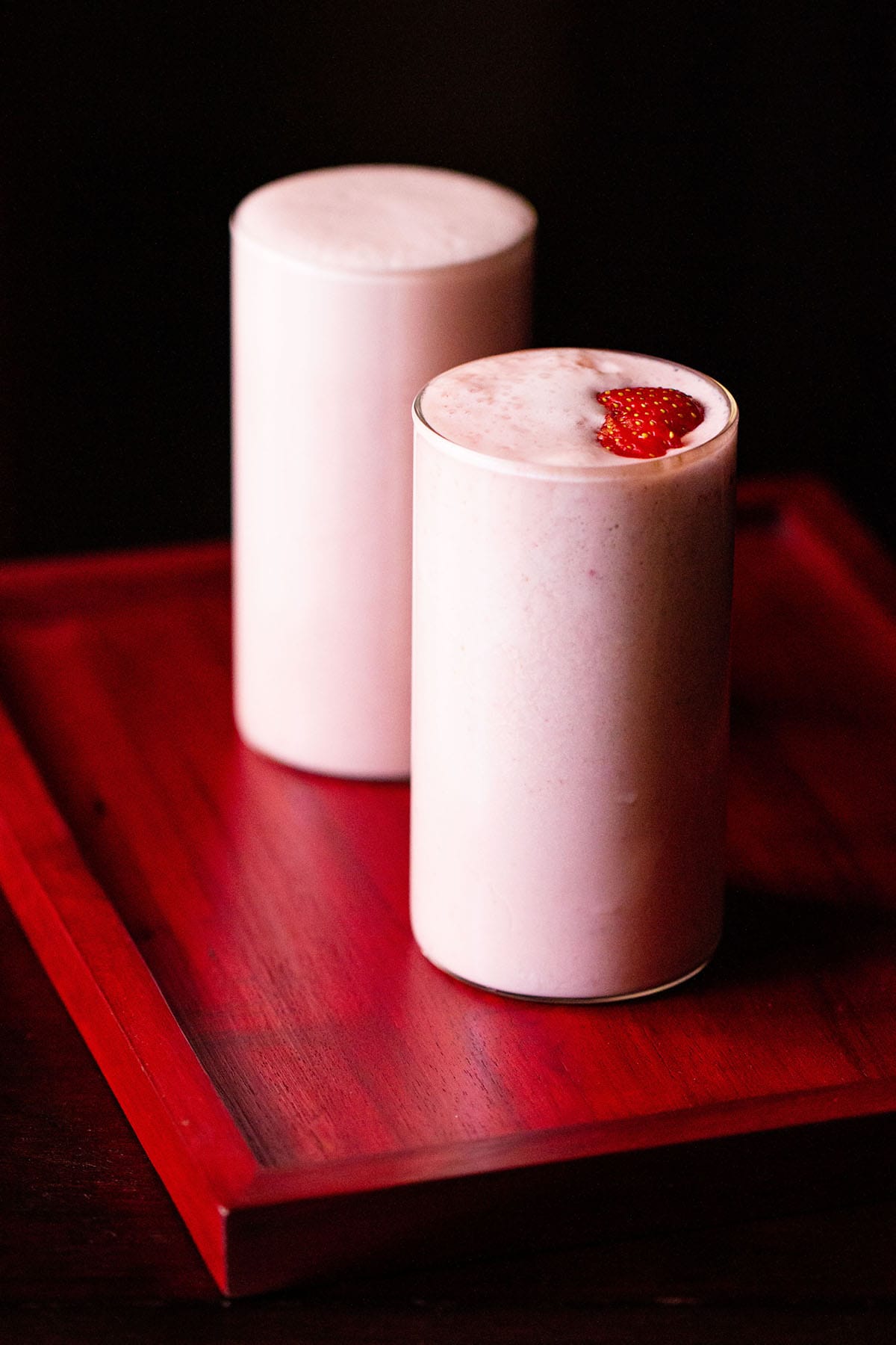 batido de fresa cubierto con rodajas de fresa en dos vasos en una bandeja de color rosa rojizo