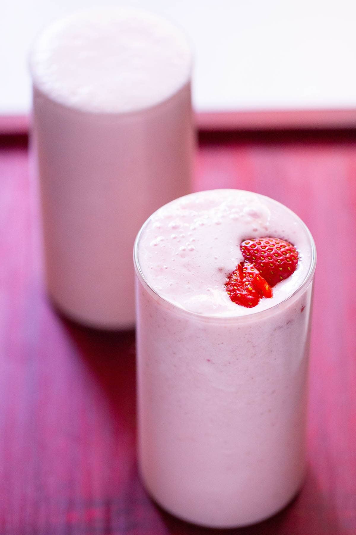 strawberry milkshake topped with strawberry slices in two glasses on a reddish pink tray