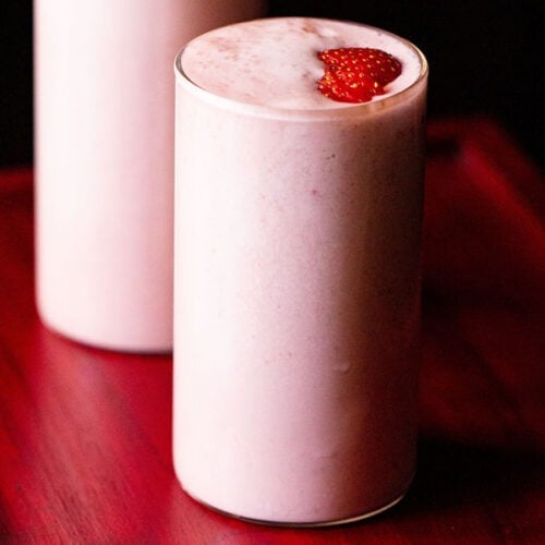 batido de fresa cubierto con rodajas de fresa en 2 vasos en una bandeja de color rosa rojizo.