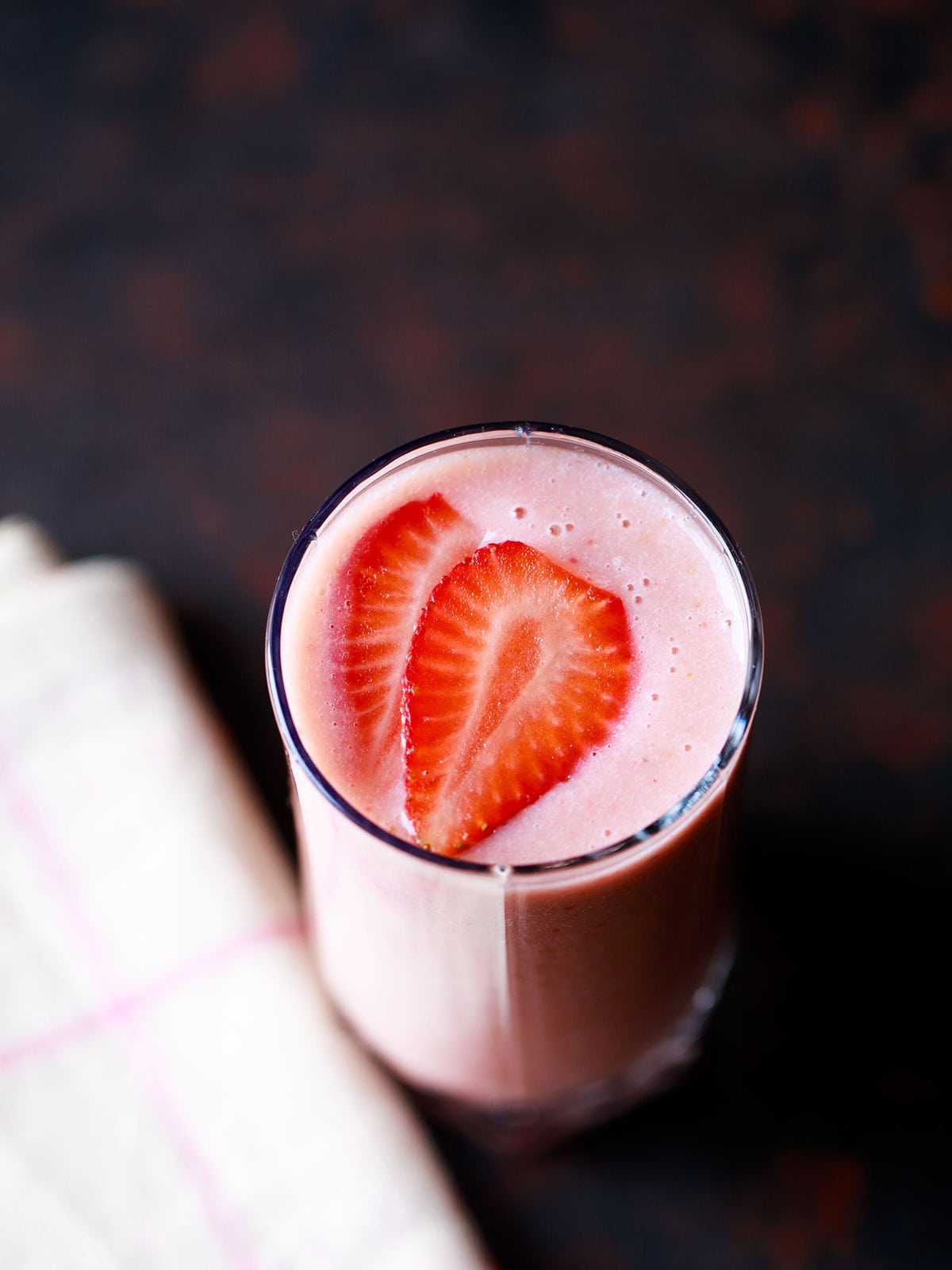 strawberry smoothie in a glass topped with strawberry slices with a cream napkin on a dark black-brown board