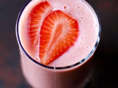 closeup overhead shot of strawberry smoothie in a glass topped with strawberry slices on a dark black-brown board