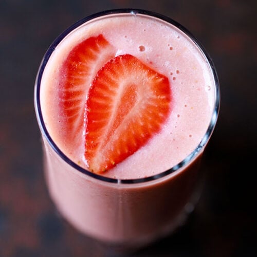 closeup overhead shot of strawberry smoothie in a glass topped with strawberry slices on a dark black-brown board