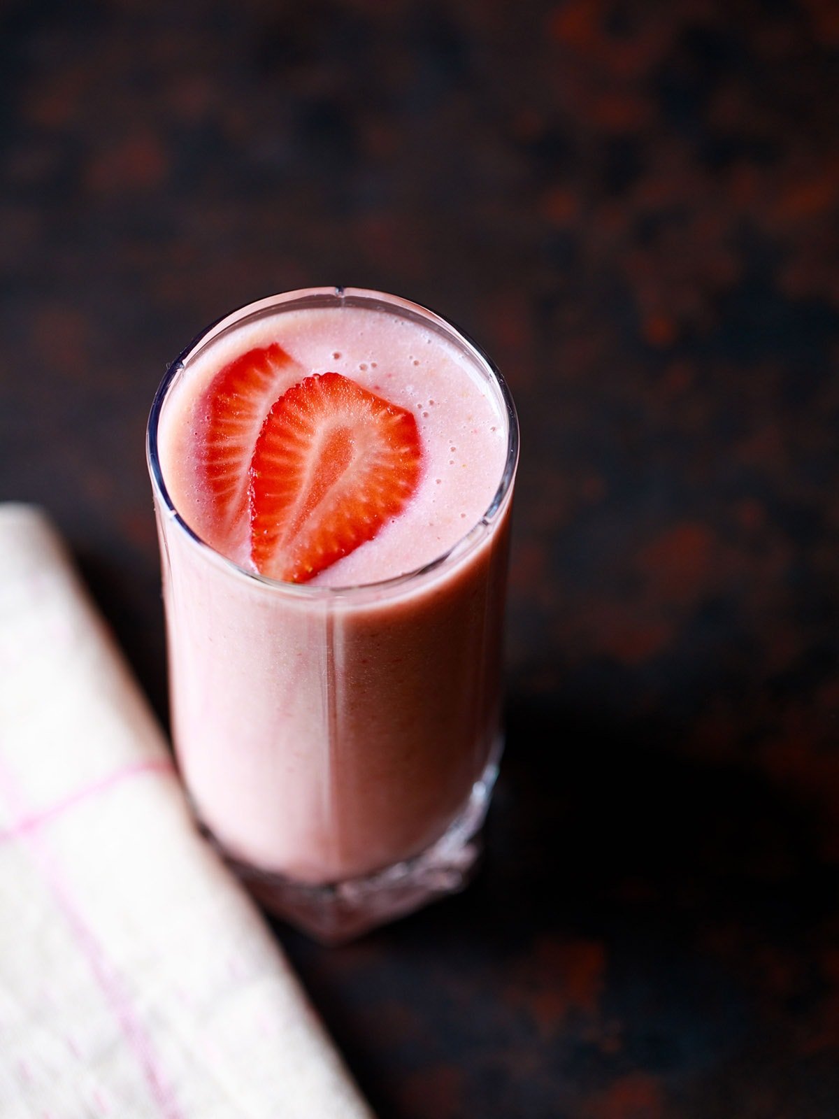 strawberry smoothie in a glass topped with strawberry slices with a cream napkin on a dark black-brown board