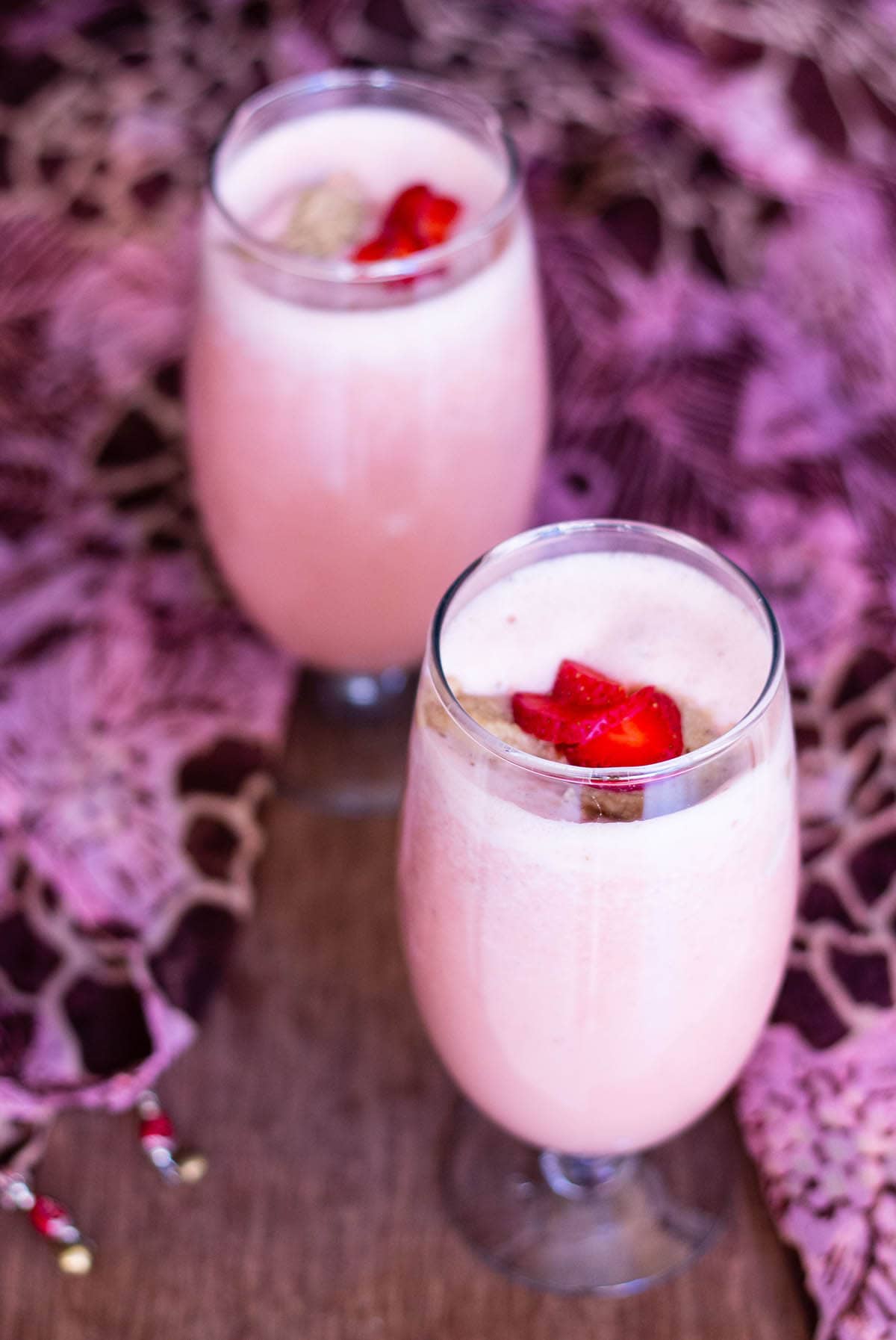 two glasses of vegan strawberry shake topped with strawberry slices and ice cream with a pink and brown fabric around on a wooden board