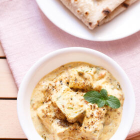 overhead shot of achari paneer in a white bowl garnished with a mint sprig on a light pink napkin with a plate of folded rotis above