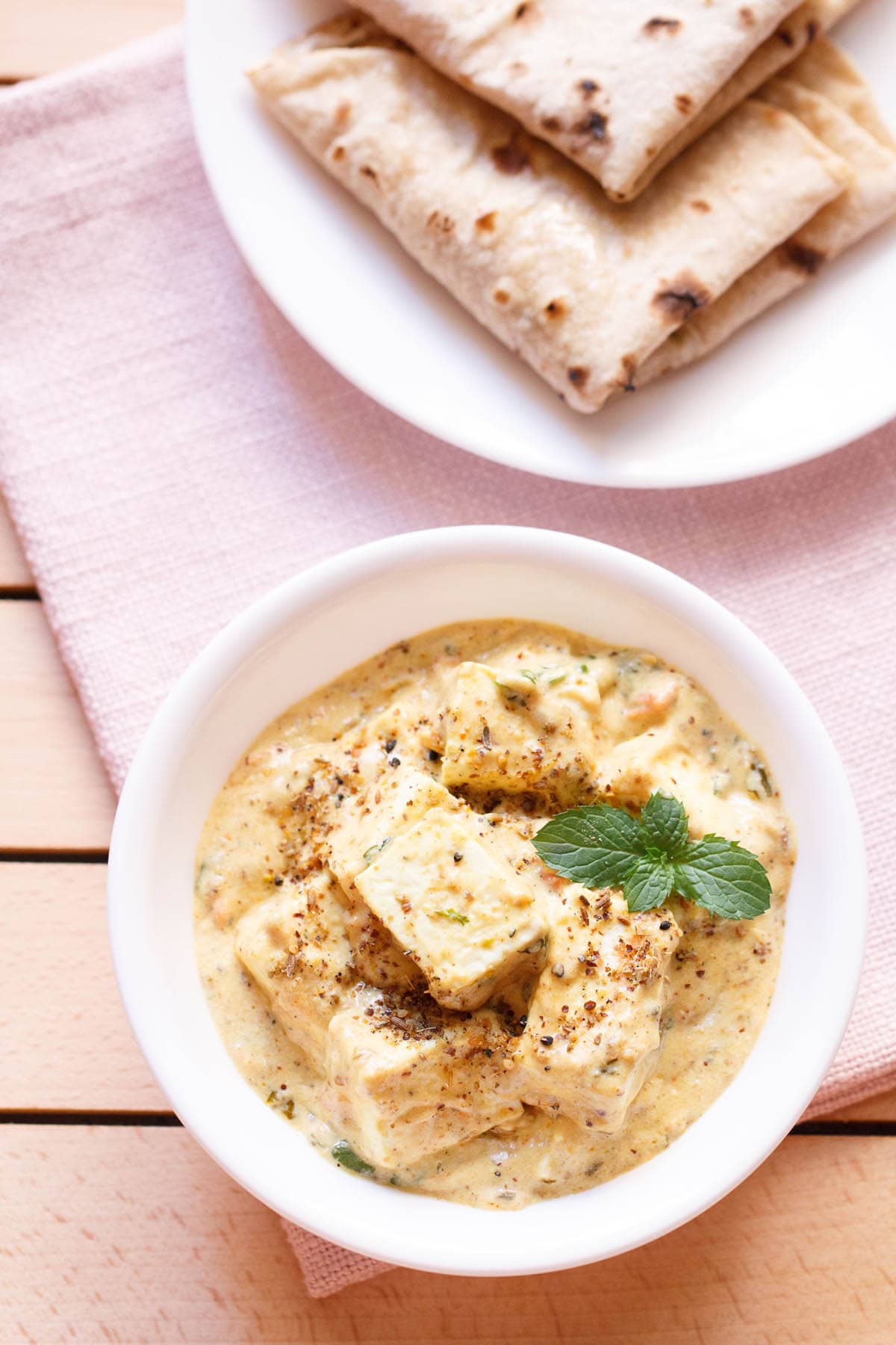 overhead shot of achari paneer in a white bowl garnished with a mint sprig on a light pink napkin with a plate of folded rotis above