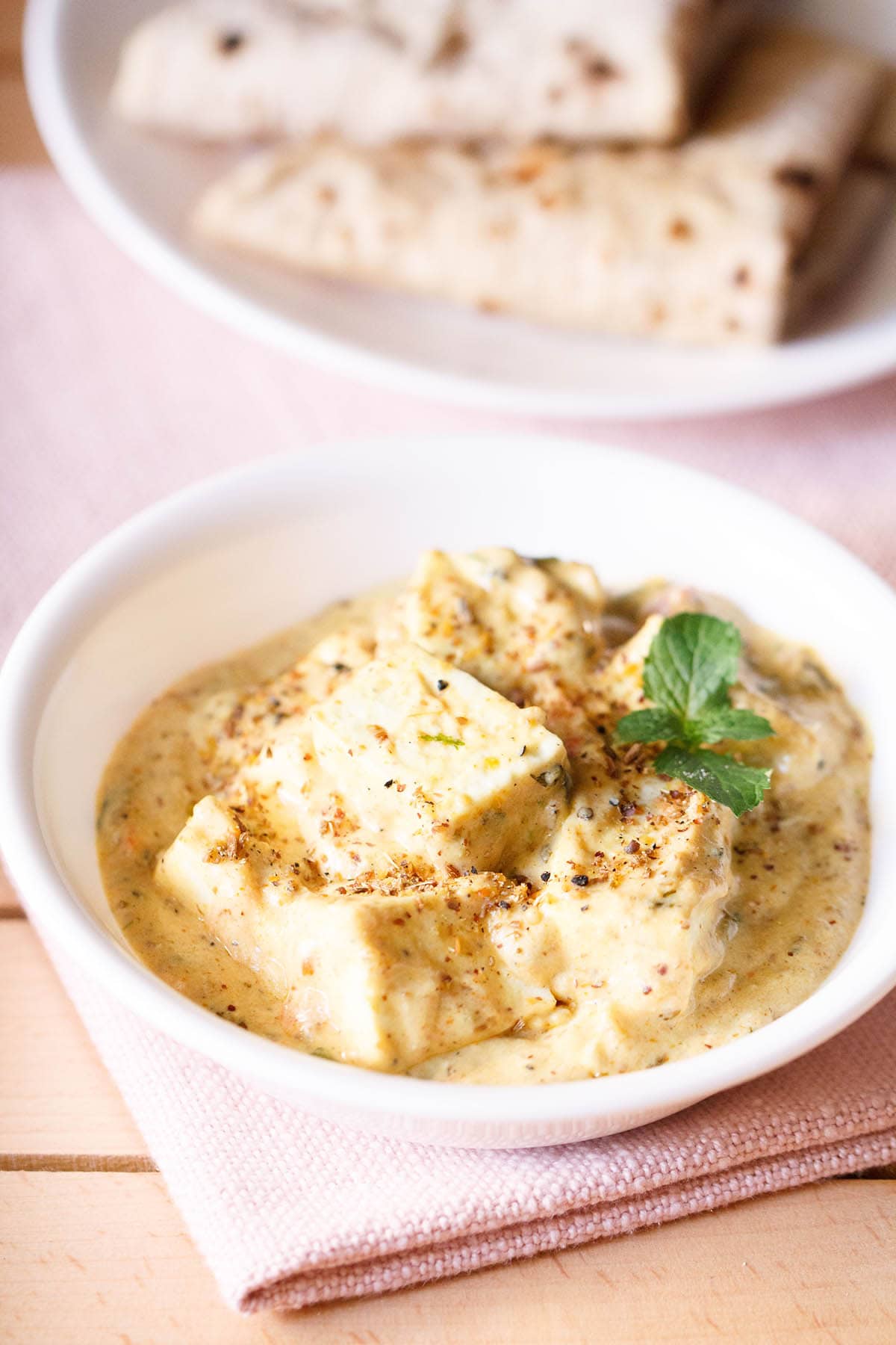 achari paneer in a white bowl garnished with a mint sprig on a light pink napkin with a plate of folded rotis above