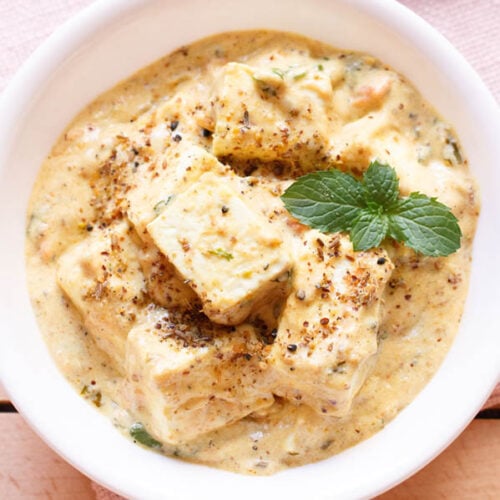 overhead shot of achari paneer in a white bowl garnished with a mint sprig on a light pink napkin