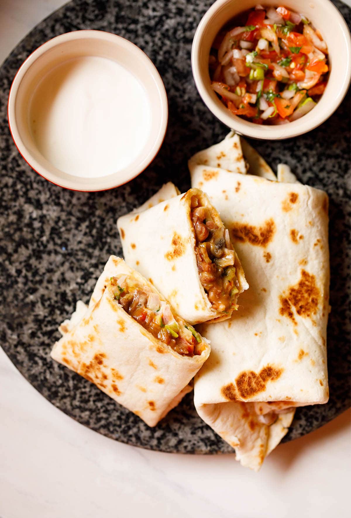 overhead shot of burrito wraps placed on a granite board with bowls filled with sour cream and guacamole on top