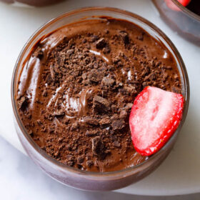 overhead shot of chocolate mousse topped with grated chocolate and a strawberry halve in a glass bowl on a white marble board