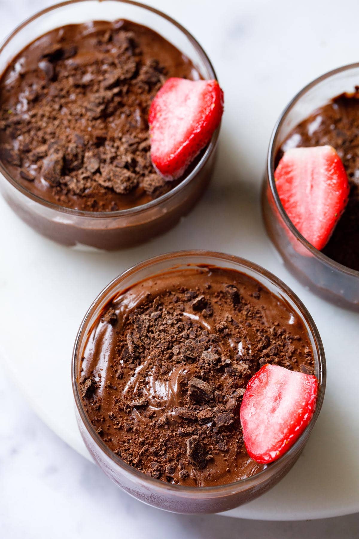 overhead shot of three bowls of chocolate mousse topped with grated chocolate and a strawberry halve in a glass bowl on a white marble board