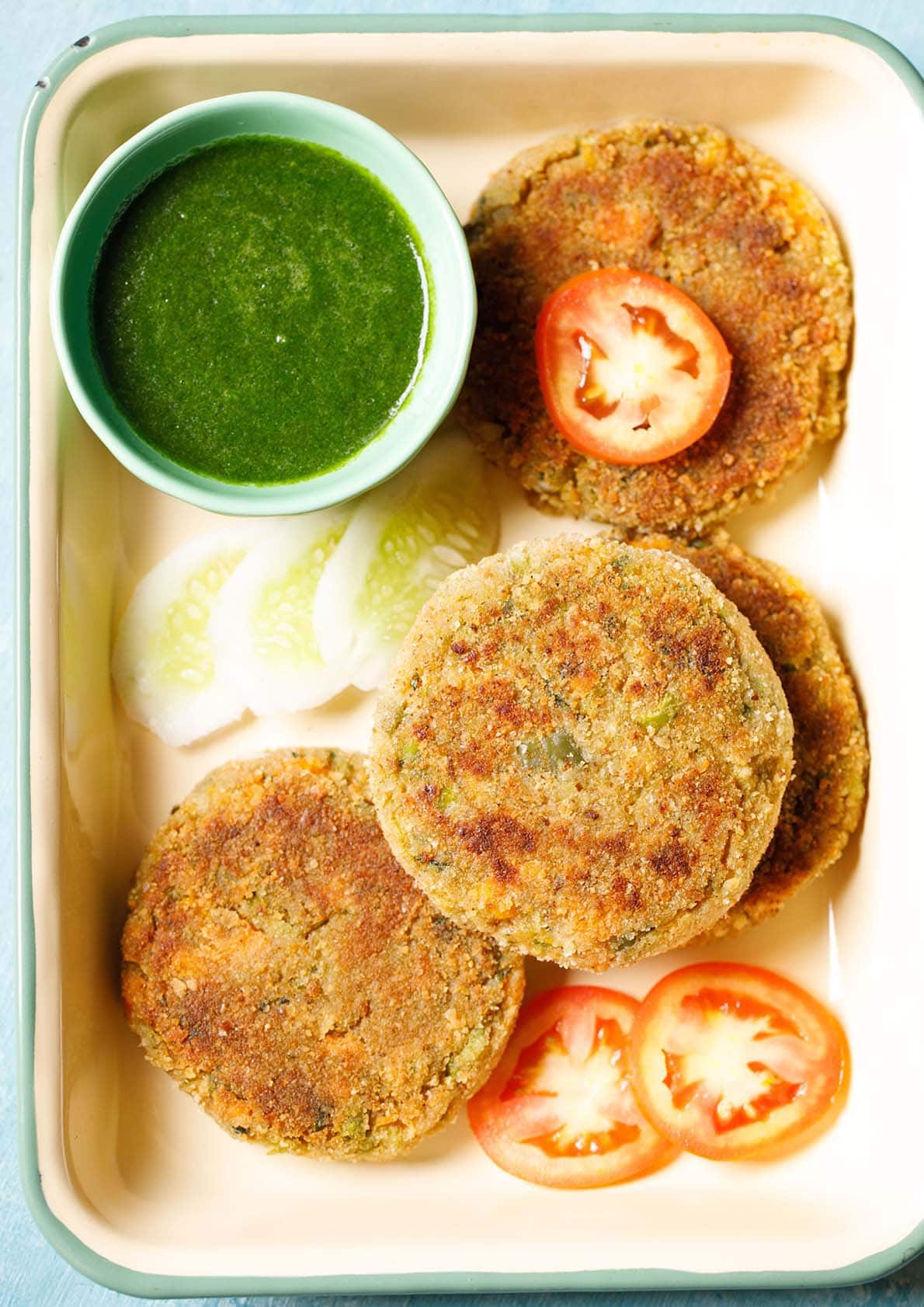 veg cutlet placed on a tray with a few slices of tomatoes, cucumber and a light green bowl filled with green chutney