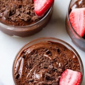 overhead shot of three bowls of chocolate mousse topped with grated chocolate and a strawberry halve in a glass bowl on a white marble board with text layovers