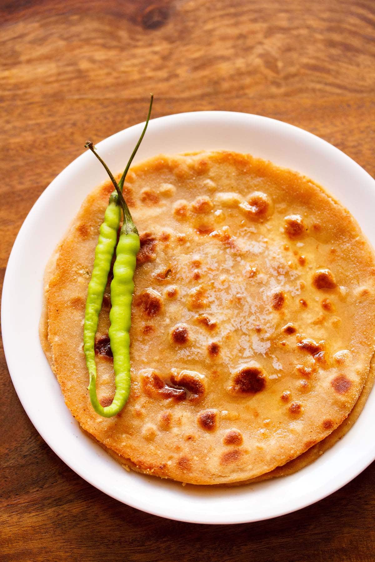 overhead shot of gobi paratha in a white plate topped with two green chillies at the side