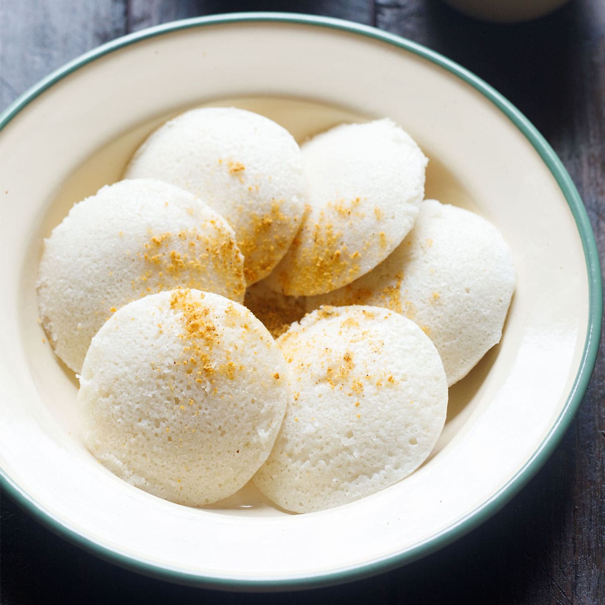 idli arranged in a cream plate sprinkled with idli podi powder on top on a dark wooden board