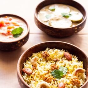 Kashmiri pulao in a brown bowl garnished with fried nuts and onions and a few fresh mint leaves next to a bowl of tomato raita on a beige table cloth