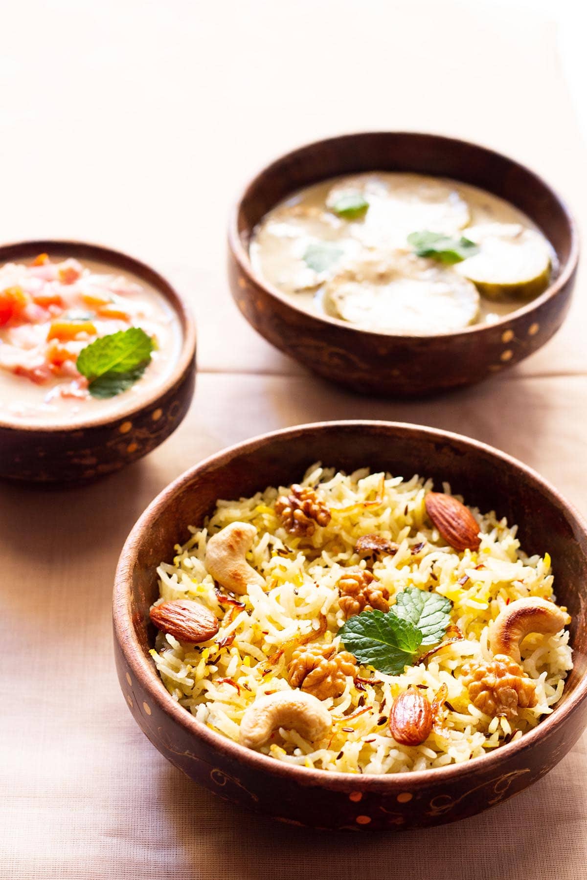 Kashmiri pulao in a brown bowl garnished with fried nuts and onions and a few fresh mint leaves next to a bowl of tomato raita on a beige table cloth