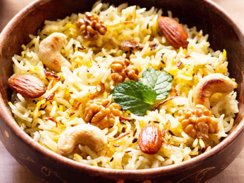 Kashmiri pulao in a brown bowl garnished with fried nuts and onions and a few fresh mint leaves next to a bowl of tomato raita on a beige table cloth