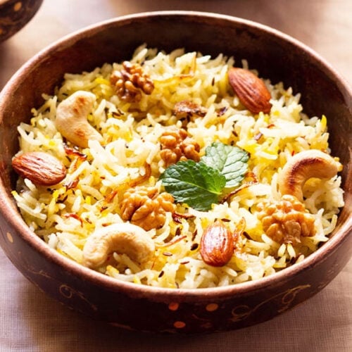 Kashmiri pulao in a brown bowl garnished with fried nuts and onions and a few fresh mint leaves next to a bowl of tomato raita on a beige table cloth