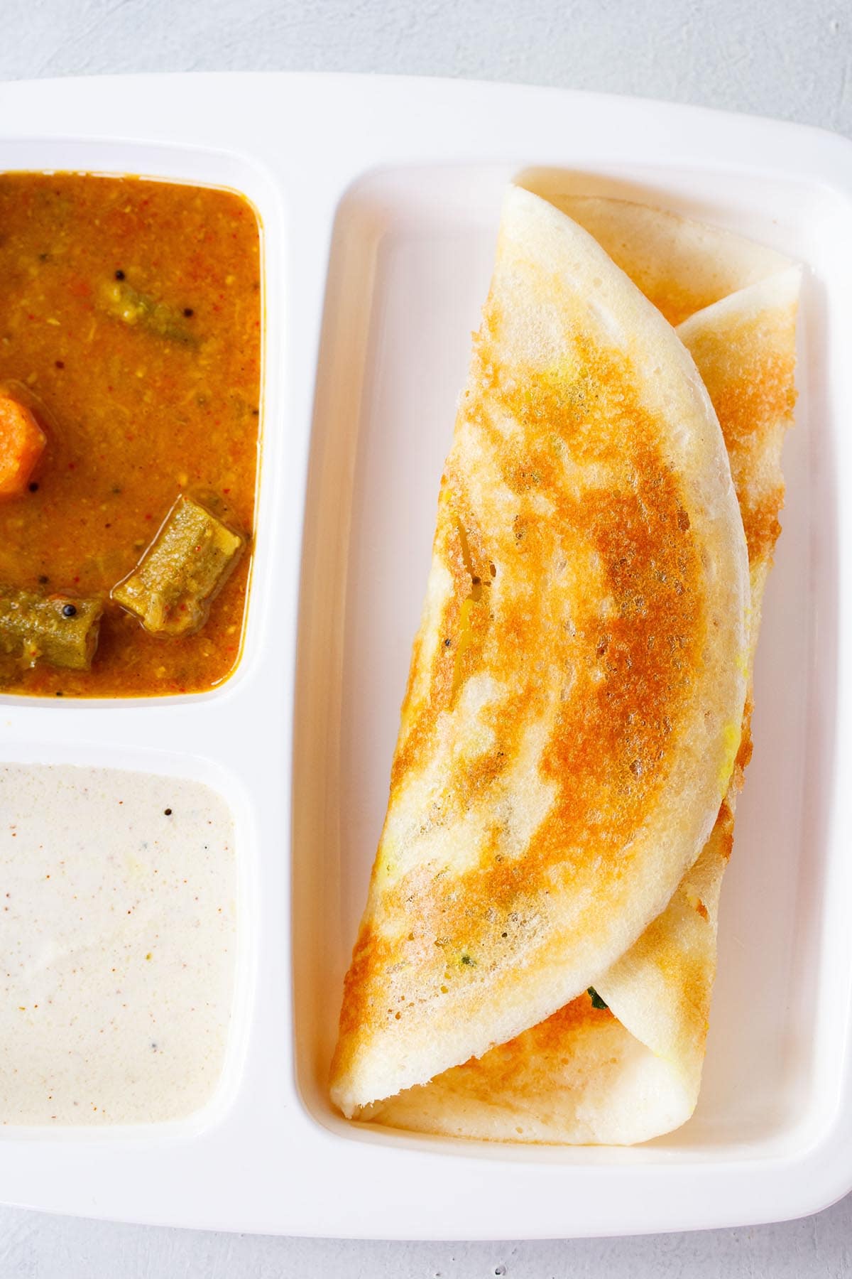 folded masala dosa in a white partitioned tray with sambar and coconut chutney