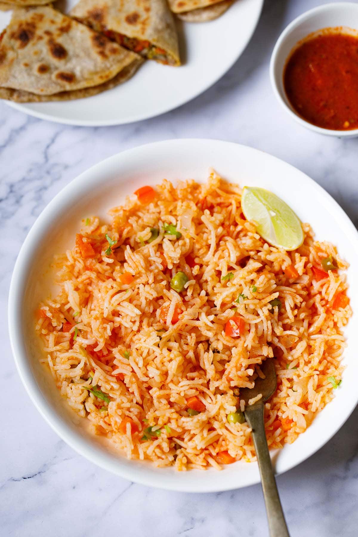 overhead shot of mexican rice with a lemon wedge and a brass fork in the rice in a large white bowl with enchilada sauce and quesadilla on top