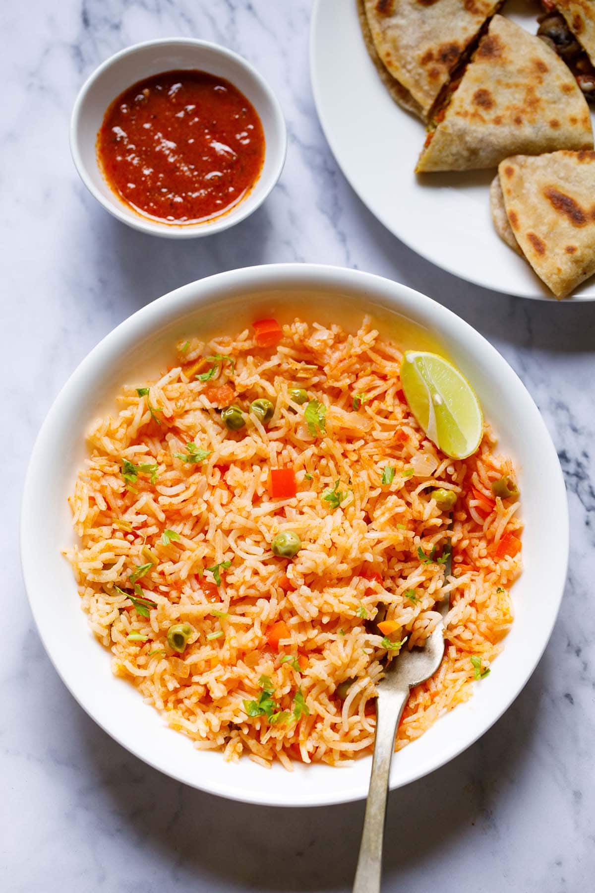 overhead shot of mexican rice with a lemon wedge and a brass fork in the rice in a large white bowl with enchilada sauce and quesadilla on top