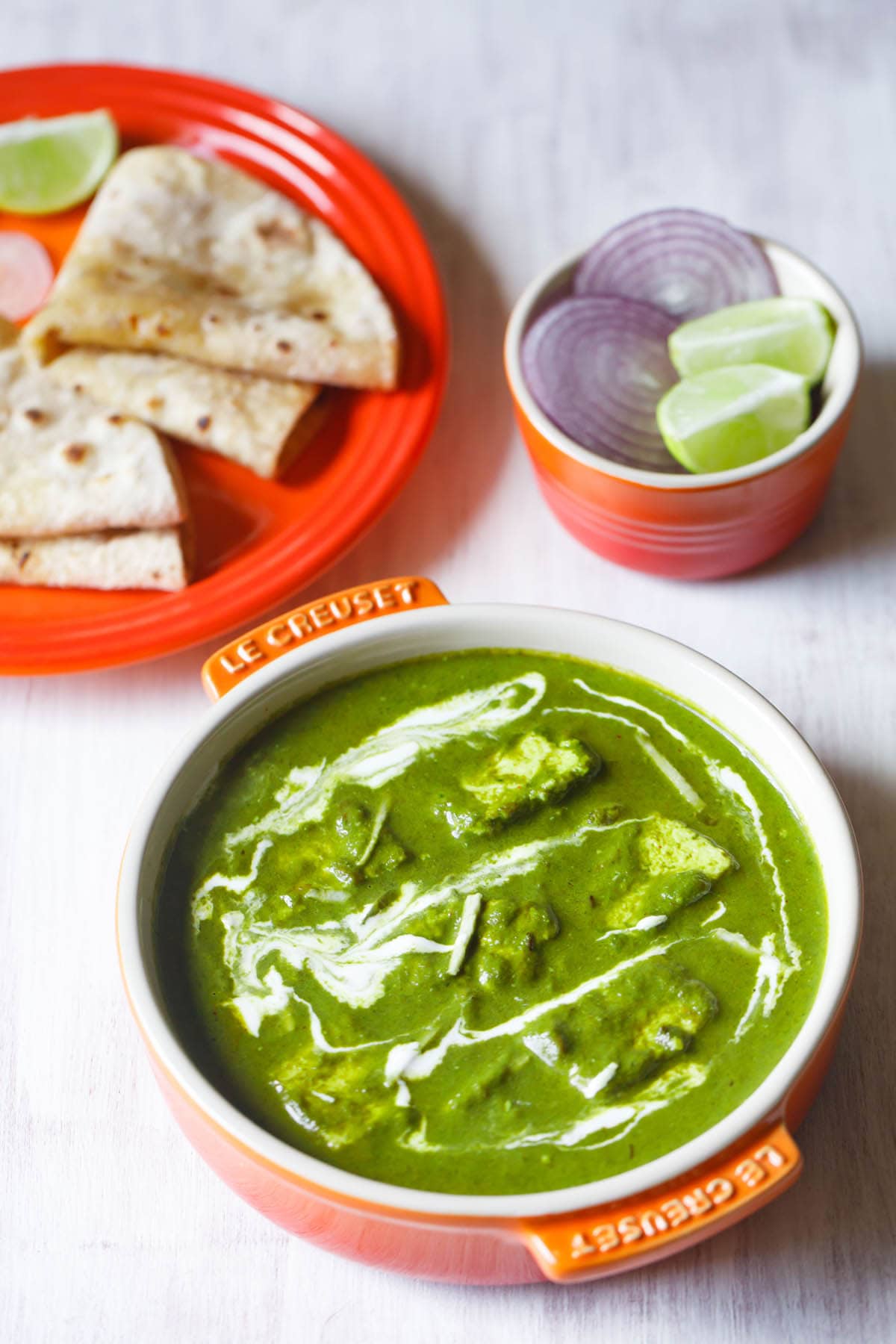 palak paneer topped with cream, garnished with ginger julienne and served in an orange bowl on a white board with roti kept on a dark orange plate on top left and a small orange bowl with round sliced onions and lemon wedges on top