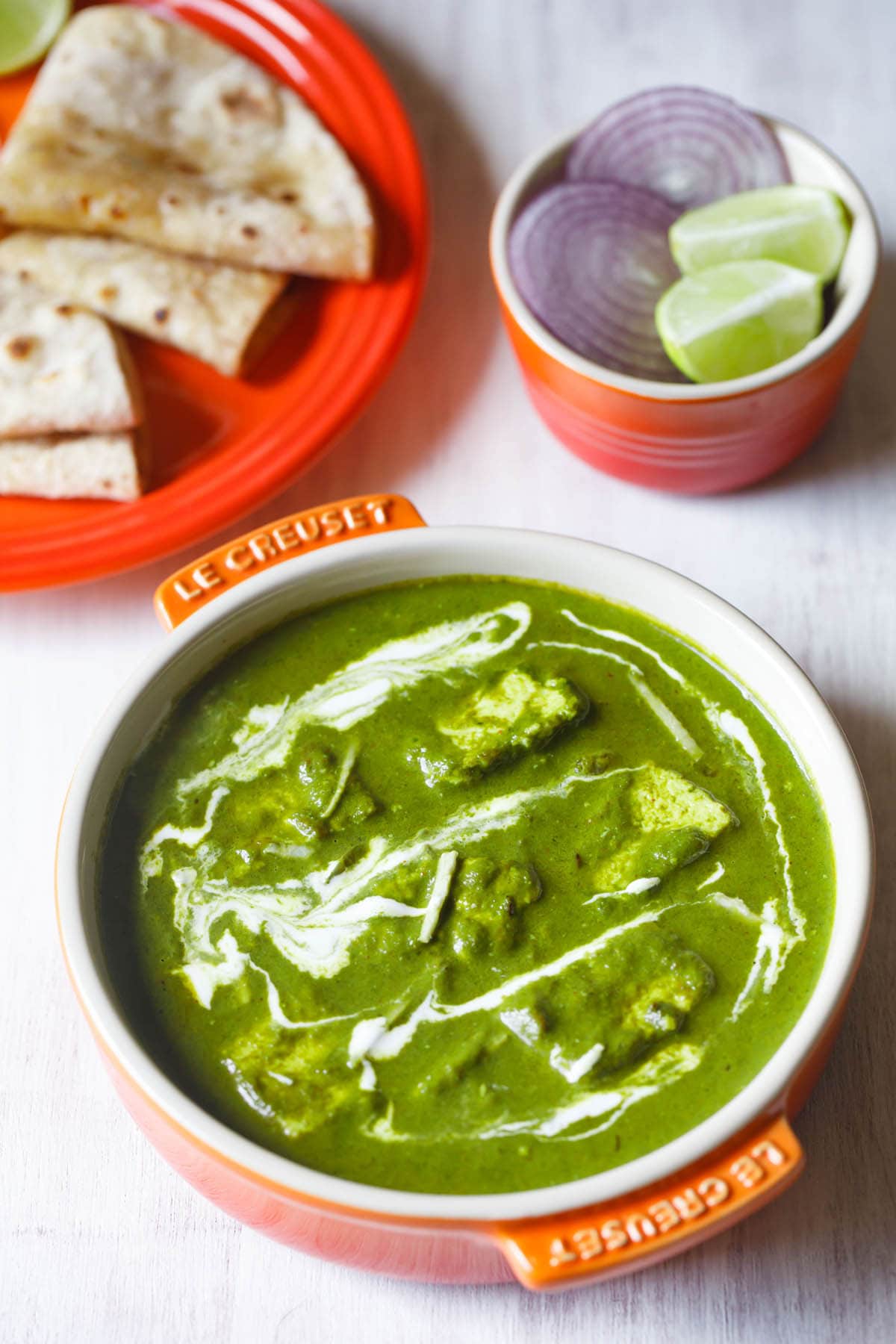 palak paneer topped with cream, garnished with ginger julienne and served in an orange bowl on a white board with roti kept on a dark orange plate on top left and a small orange bowl with round sliced onions and lemon wedges on top