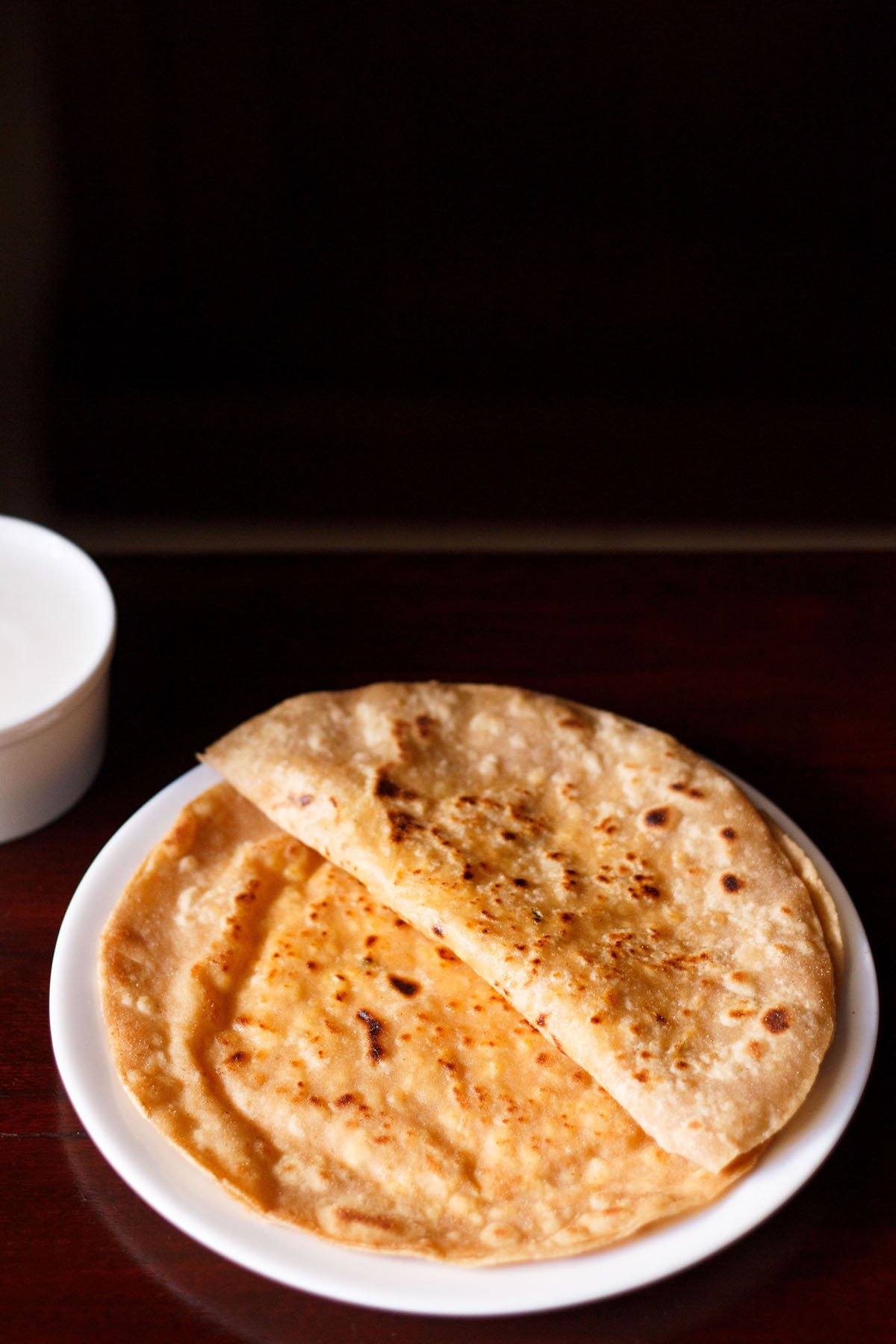 folded paneer paratha on top of a paneer paratha on a white plate on a dark brown table