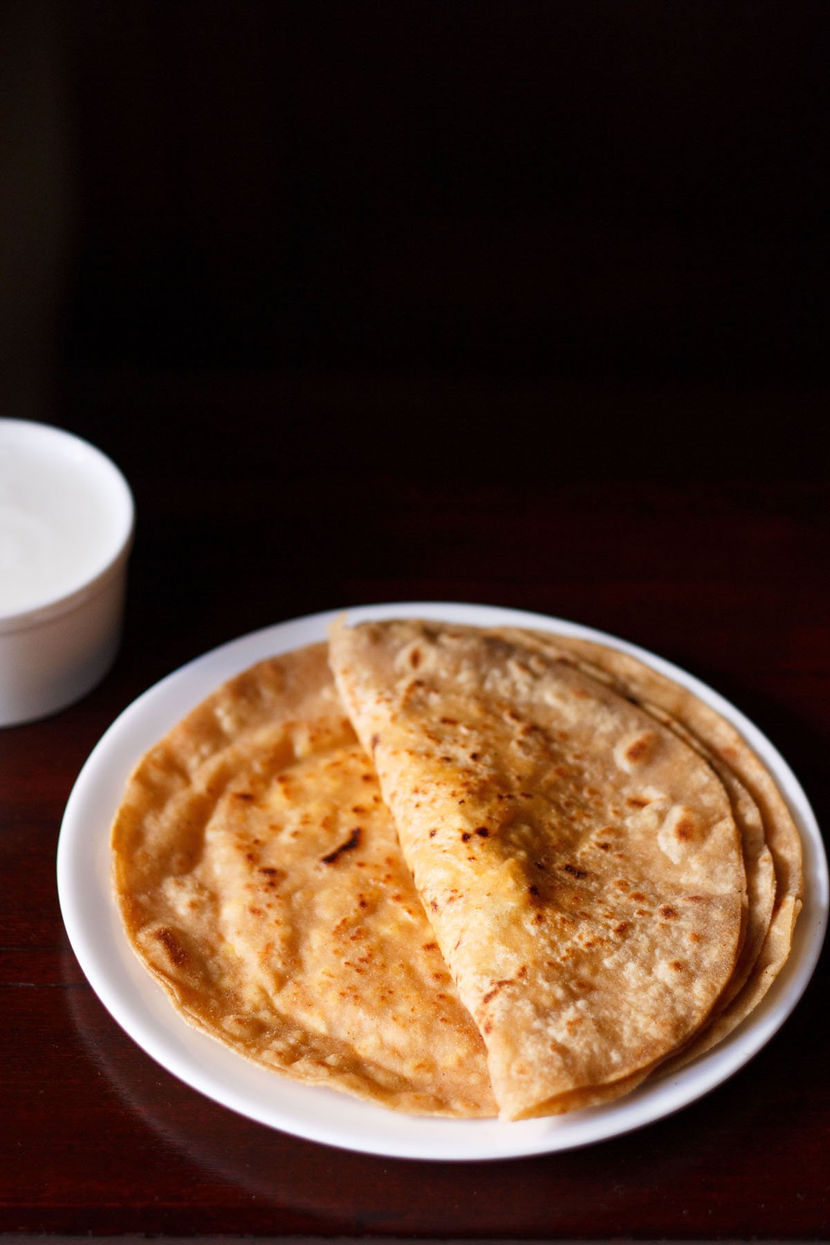 folded paneer paratha on top of another paratha on a white plate on a dark brown table.