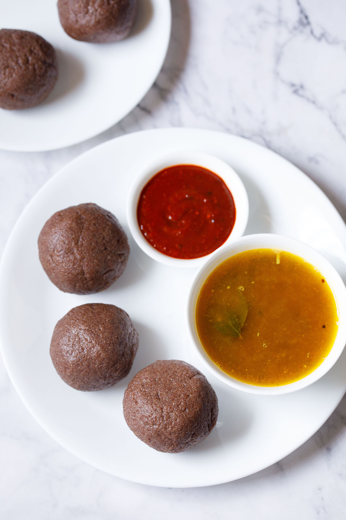 overhead shot of 3 ragi mudde balls on a white plate with two bowls of saaru and red chilli chutney
