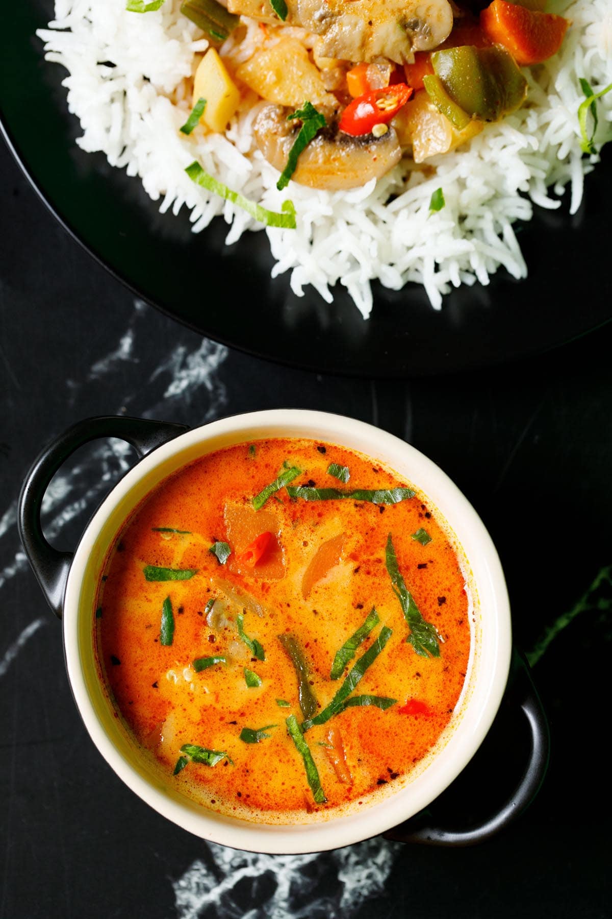 overhead shot of thai curry filled in a cream bowl with a black plate of rice and curry on top on black marble table