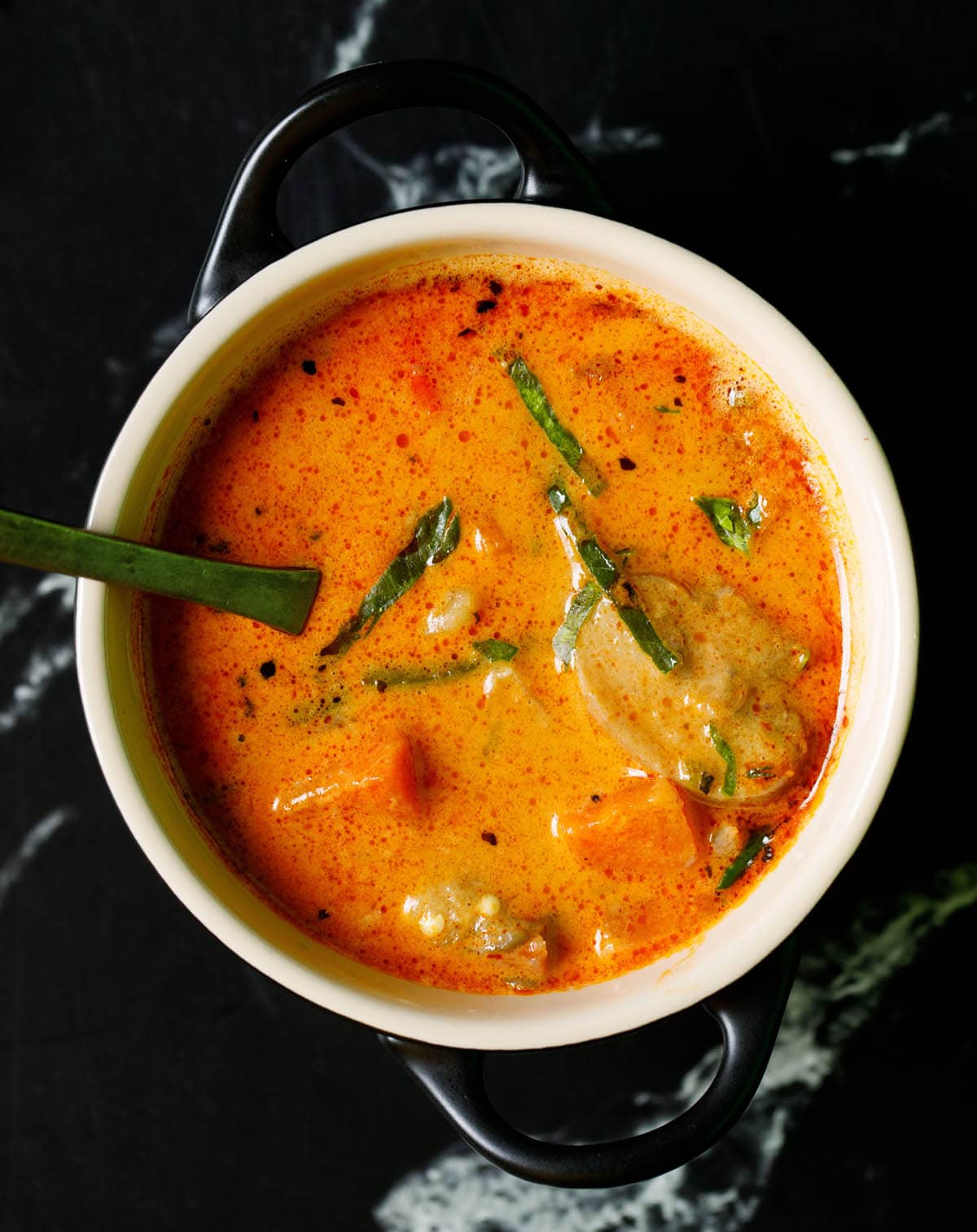 overhead shot of thai red curry filled in a cream bowl with spoon inside on a black marble table