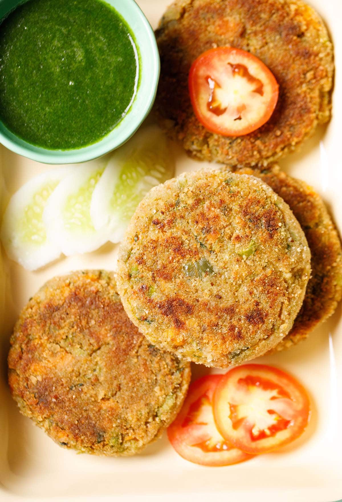cutlets placed on a tray with a few slices of tomatoes, cucumber and a light green bowl filled with green chutney