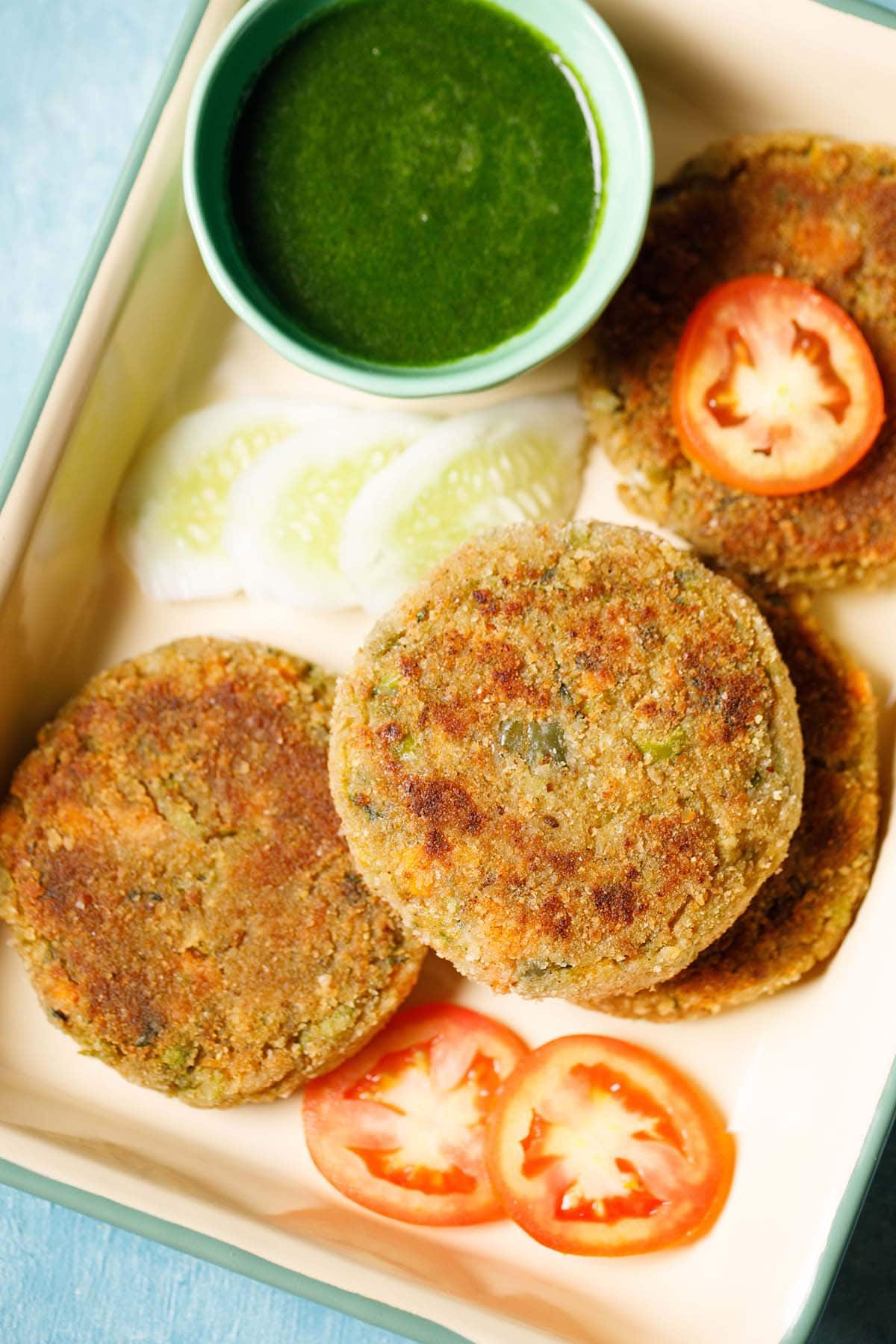 closeup shot of veg cutlet cutlet placed on a tray with a few slices of tomatoes, cucumber and a light green bowl filled with green chutney.