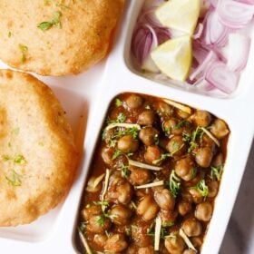 closeup shot of white plate with sections filled with chana masala, sliced onions, fresh lime wedges and bhatura on a marble table-top