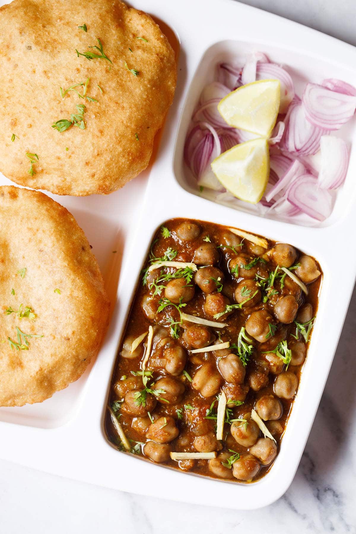 closeup shot of white plate with sections filled with chana masala, sliced onions, fresh lime wedges and bhatura on a marble table-top