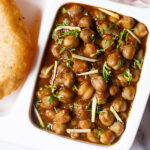 white plate with sections filled with punjabi chole masala, sliced onions, fresh lemon wedges and bhatura on a marble table-top.