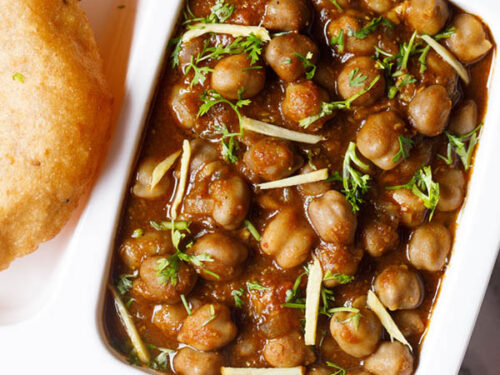 white plate with sections filled with punjabi chole masala, sliced onions, fresh lemon wedges and bhatura on a marble table-top.