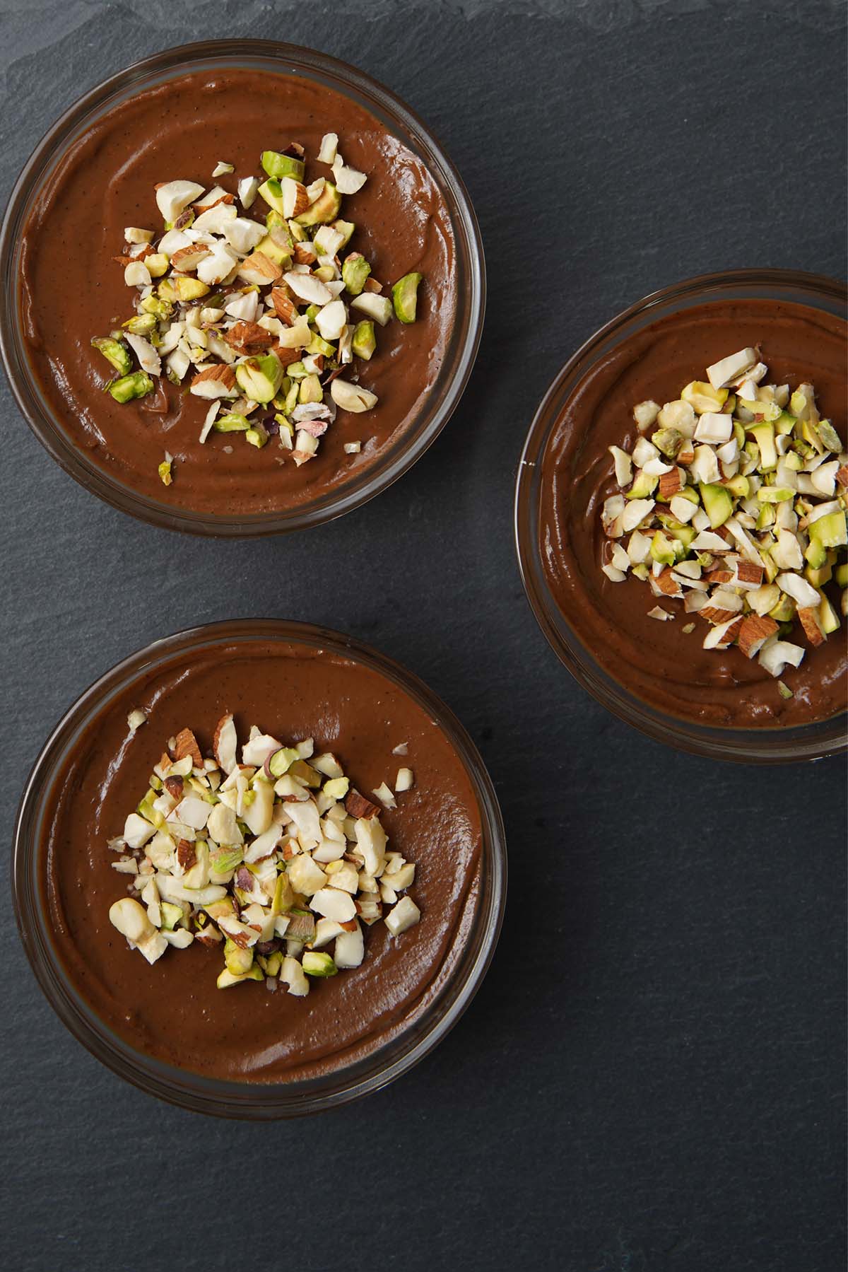 overhead shot of chocolate avocado mousse in three bowls topped with nuts on a black granite board
