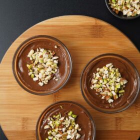 overhead shot of chocolate avocado mousse in three bowls topped with nuts on a round wooden board