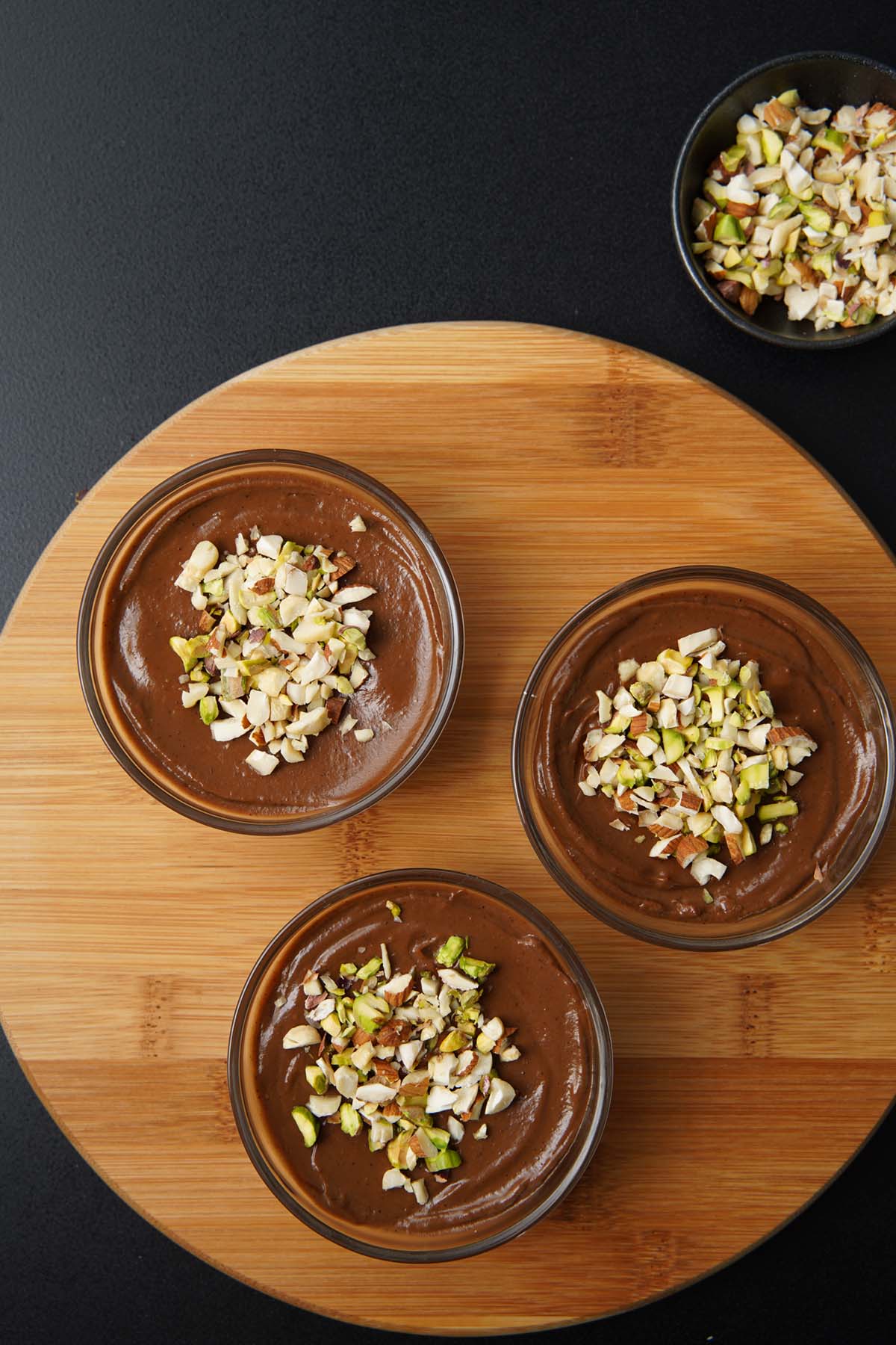 overhead shot of chocolate avocado mousse in three bowls topped with nuts on a round wooden board