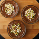 overhead shot of chocolate avocado mousse in three bowls topped with nuts on a round wooden board