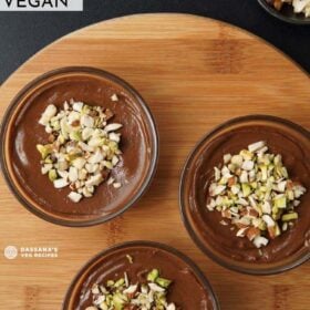 overhead shot of chocolate avocado mousse in three bowls topped with nuts on a round wooden board with text layovers